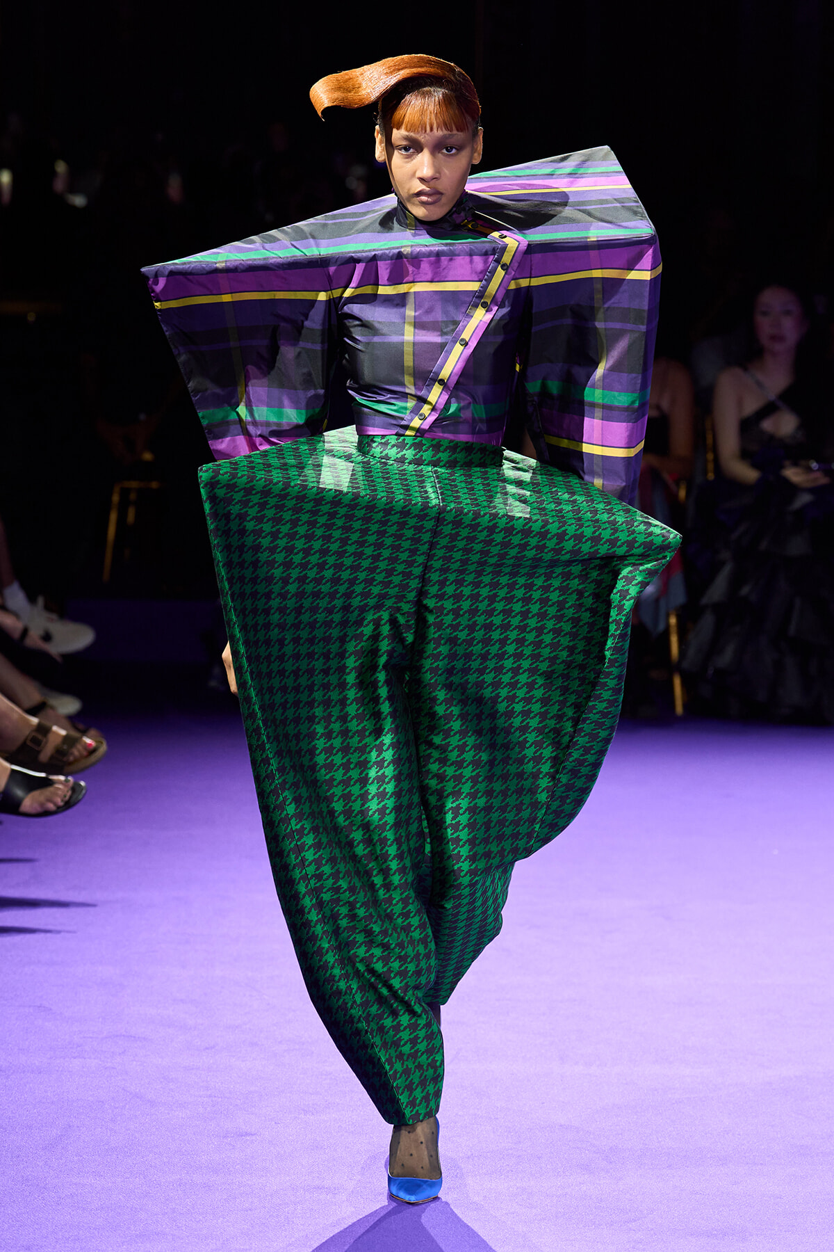 Model on a purple-lit runway wearing a sculptural purple-striped top and oversized green houndstooth pants.