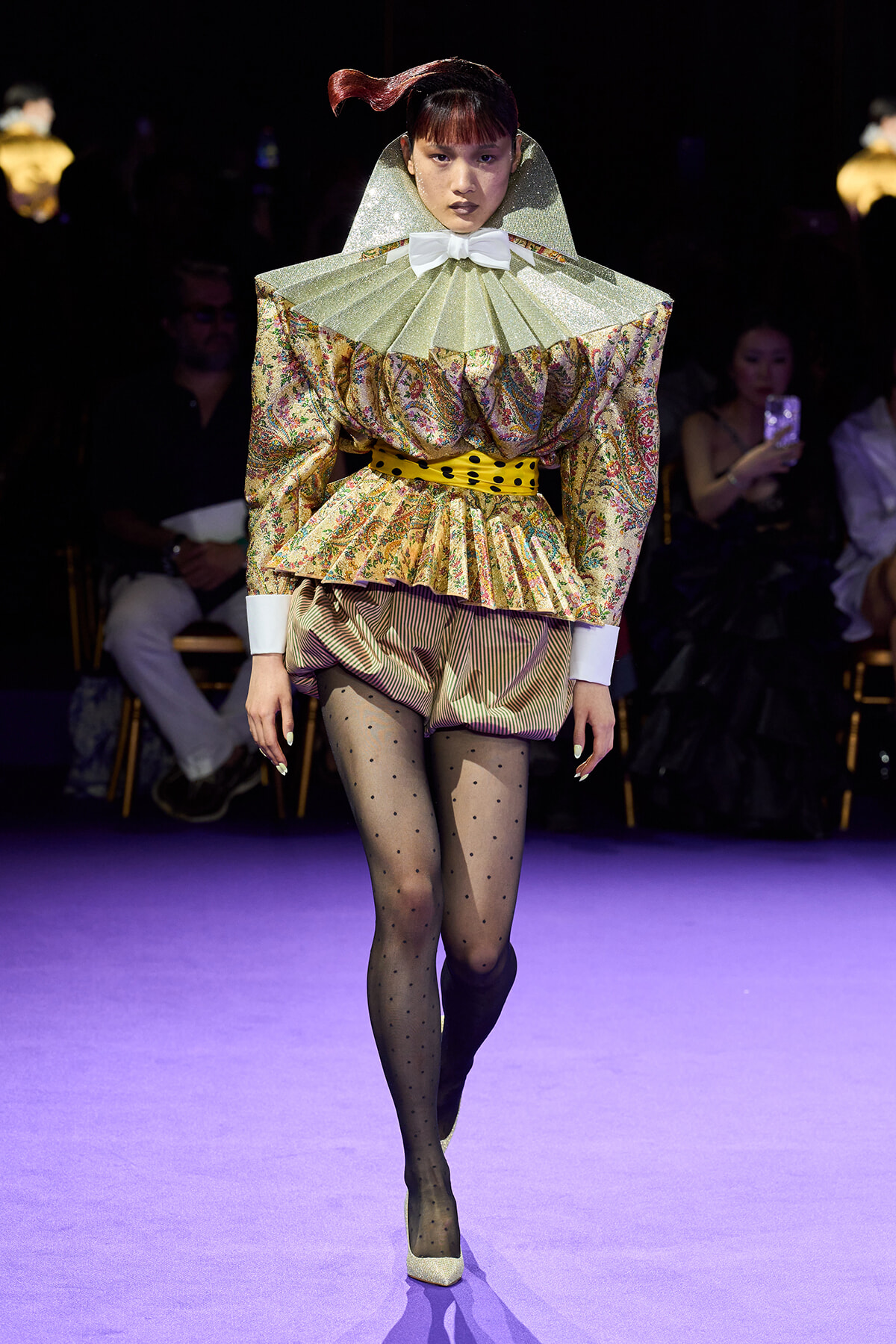 Model walking a fashion runway in a gold patterned, ruffled top with oversized green collar, white bow, yellow polka belt, and striped shorts paired with sheer dotted tights and glittery heels.