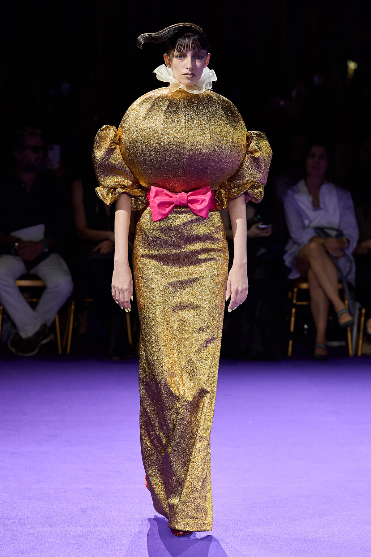 Female model walks a fashion show in a shiny gold gown with oversized puff sleeves, a white ruffled collar, and a bright pink bow at the waist.