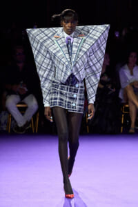 Runway model in an oversized plaid jacket with angular shoulders and a matching mini skirt, black tights, and red heels under purple lighting.