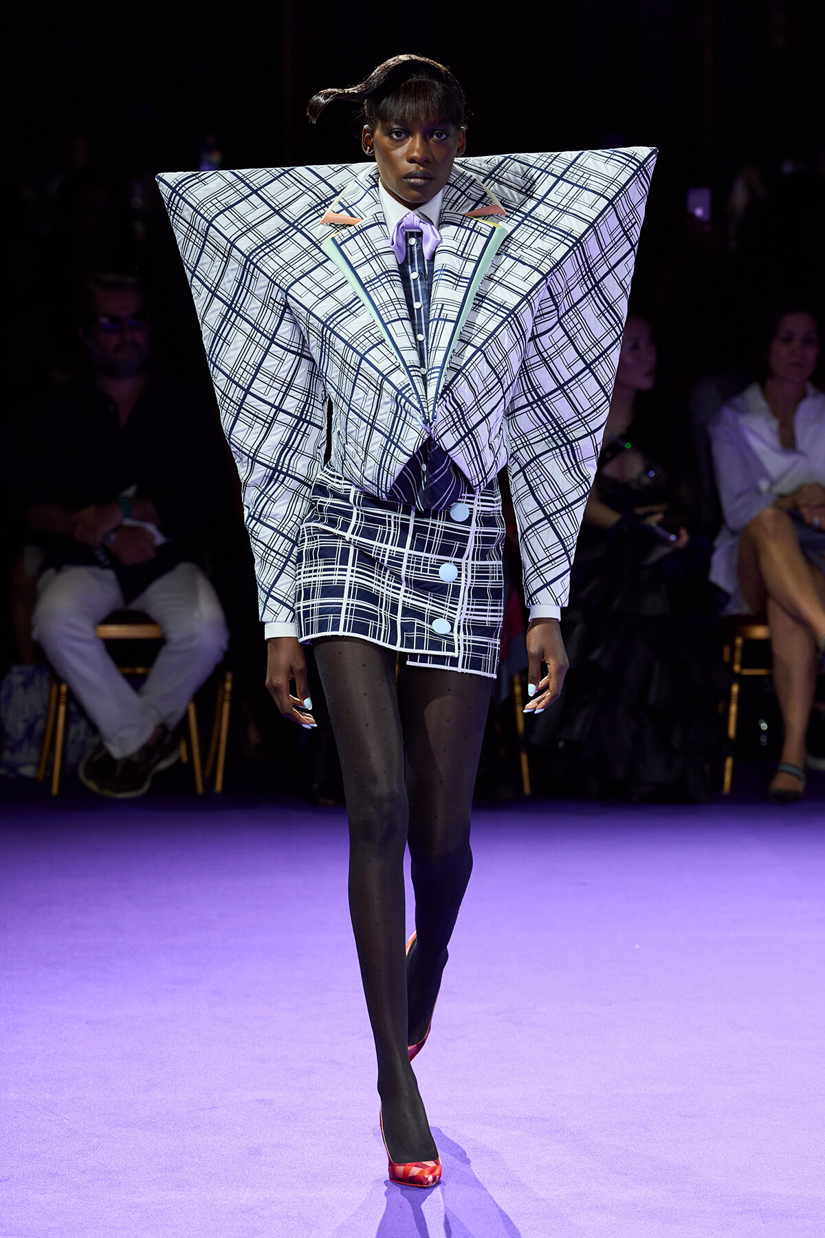 Runway model in an oversized plaid jacket with angular shoulders and a matching mini skirt, black tights, and red heels under purple lighting.