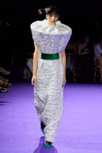 Model walks the runway in a sculptural white textured gown with oversized shoulders and a wide green belt, paired with green heels on a purple-lit catwalk.]","A fashion model wears a crinkled white dress with a ruffled collar and emerald belt, complementing bright green pointed-toe shoes."","Runway image: female model in a voluminous, crinkled white dress cinched at the waist with a green belt and green shoes on a purple-lit floor."","Editorial shot of a model in an avant-garde white gown with pronounced shoulder silhouette, green belt, and matching green footwear on a fashion show runway.