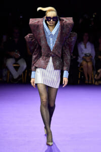 Runway model wearing a voluminous burgundy floral blazer over a blue houndstooth shirt, striped mini skirt, polka-dot tights, and colorful heels with oversized sunglasses.