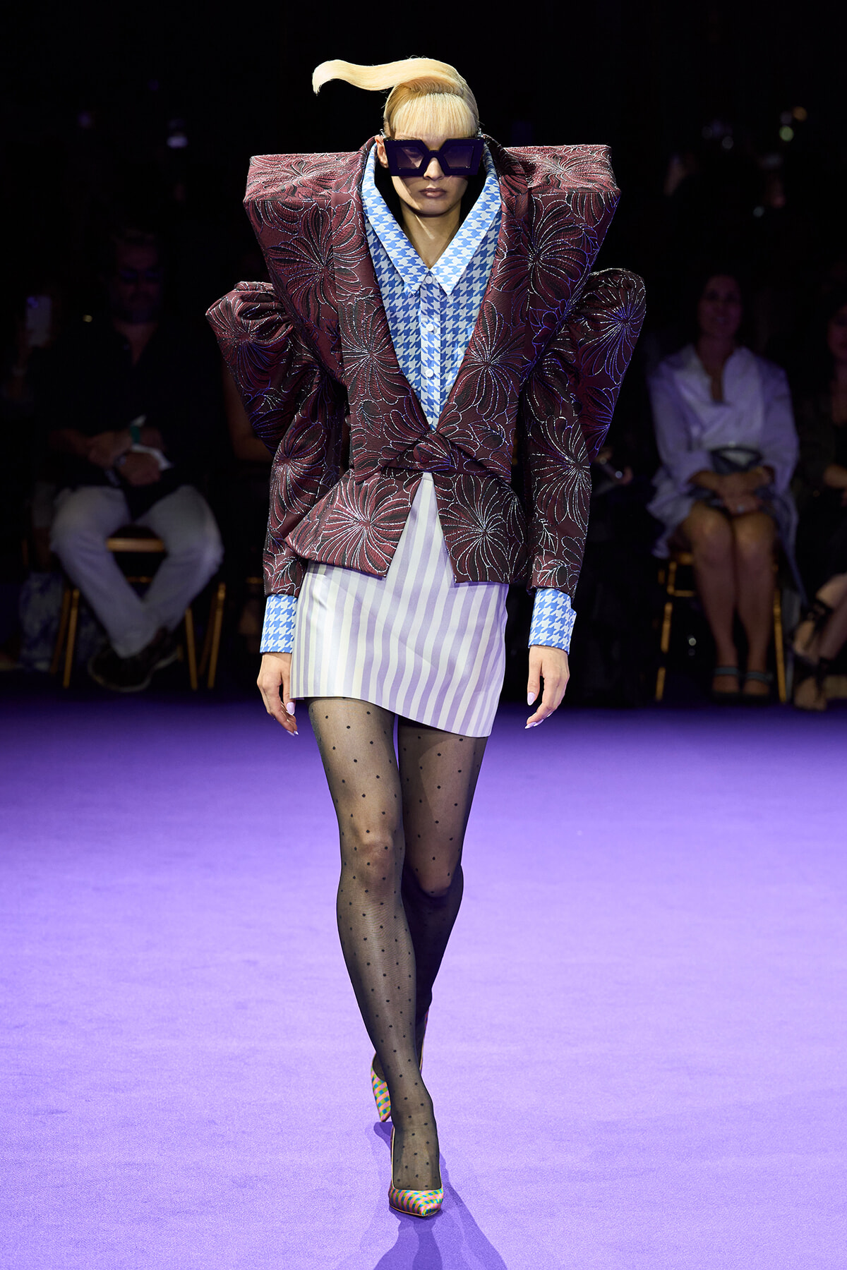 Runway model wearing a voluminous burgundy floral blazer over a blue houndstooth shirt, striped mini skirt, polka-dot tights, and colorful heels with oversized sunglasses.