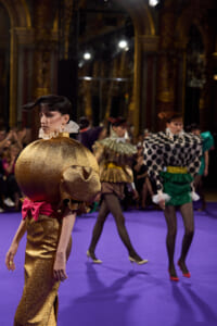 Model walks a runway in a glittering gold pumpkin-shaped dress with pink bow, others in bold patterned outfits behind her on a purple floor.