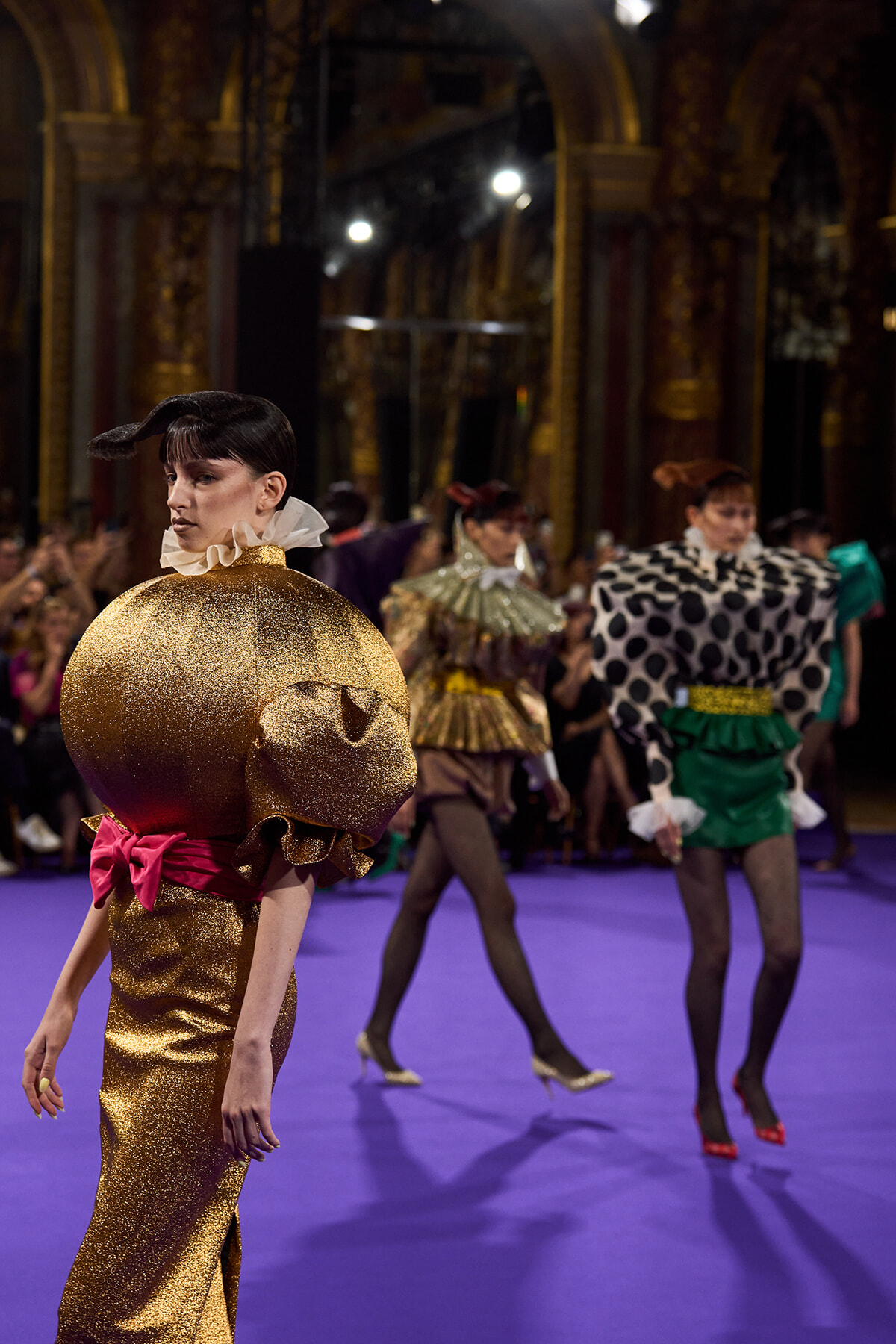 Model walks a runway in a glittering gold pumpkin-shaped dress with pink bow, others in bold patterned outfits behind her on a purple floor.