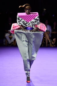 Model walking a purple-lit runway in a bold, oversized outfit: pink polka-dot and black floral wrap with large pink sleeves and metallic silver trousers.