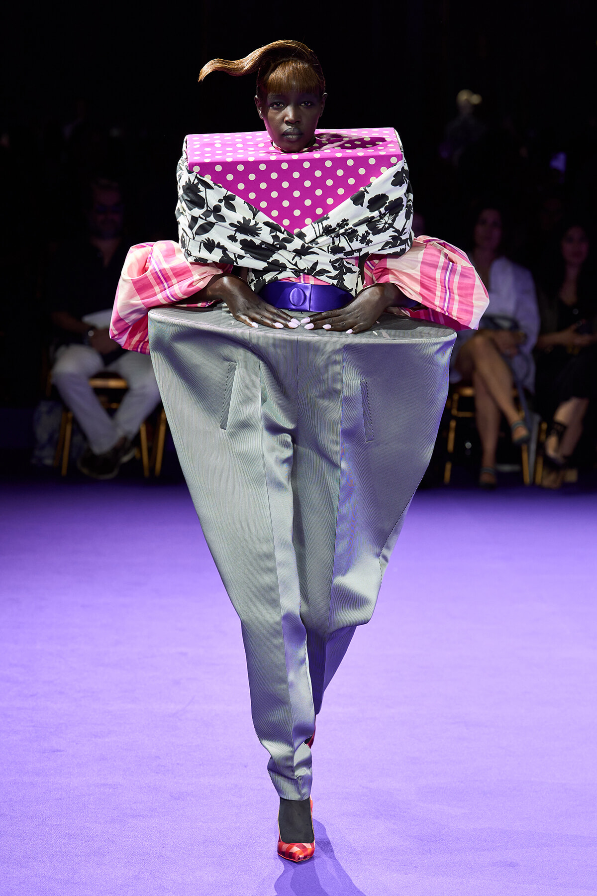 Model walking a purple-lit runway in a bold, oversized outfit: pink polka-dot and black floral wrap with large pink sleeves and metallic silver trousers.