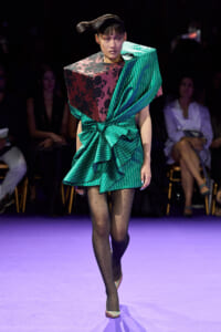 Runway model in a sculptural green striped dress with a burgundy floral bodice and a large teal drape, walking on a purple catwalk.
