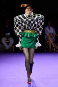Model walks runway in a bold geometric outfit: oversized black-and-white polka dot top, green mini skirt with yellow leopard belt, sheer dotted tights, and red heels.