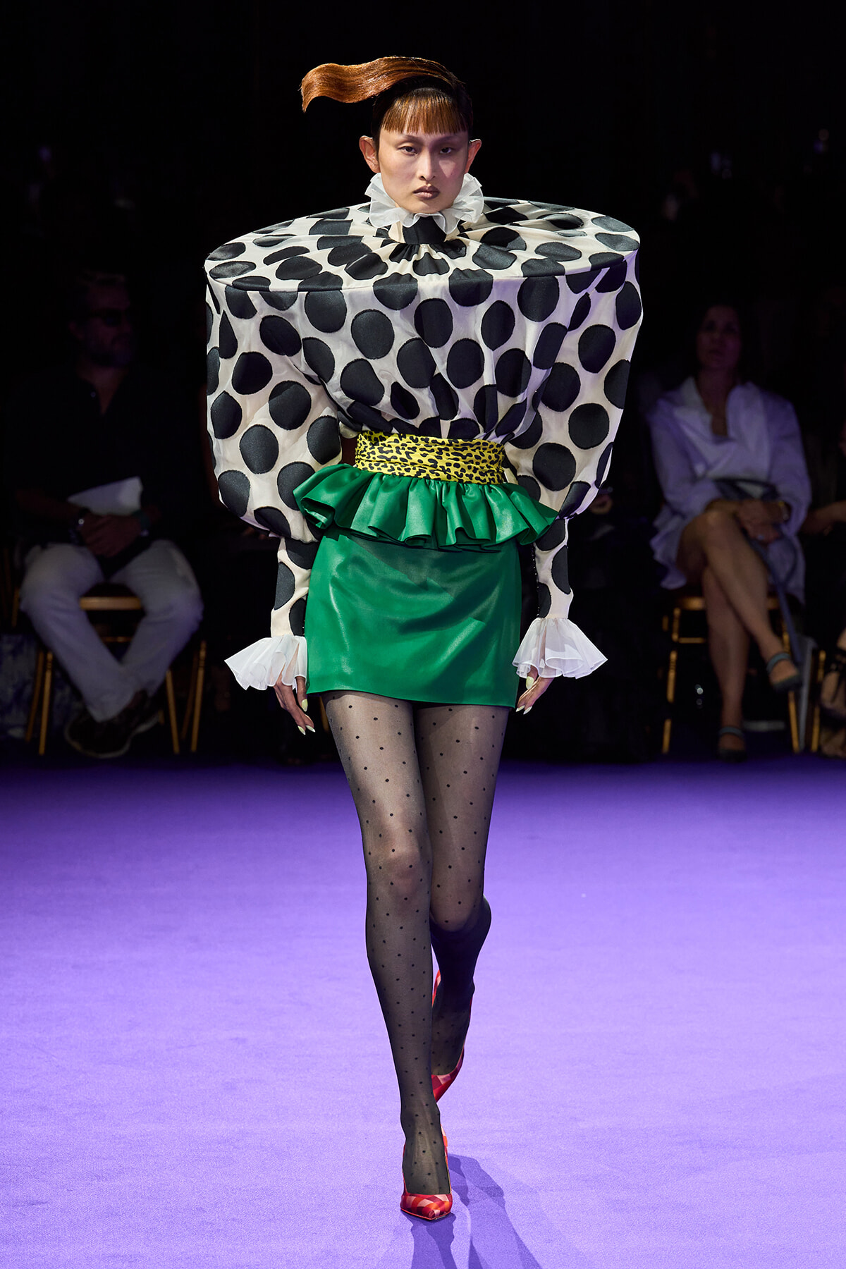 Model walks runway in a bold geometric outfit: oversized black-and-white polka dot top, green mini skirt with yellow leopard belt, sheer dotted tights, and red heels.