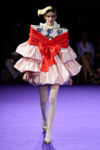 Runway model in a voluminous pink striped gown with a bold red sash bow and floral bodice.