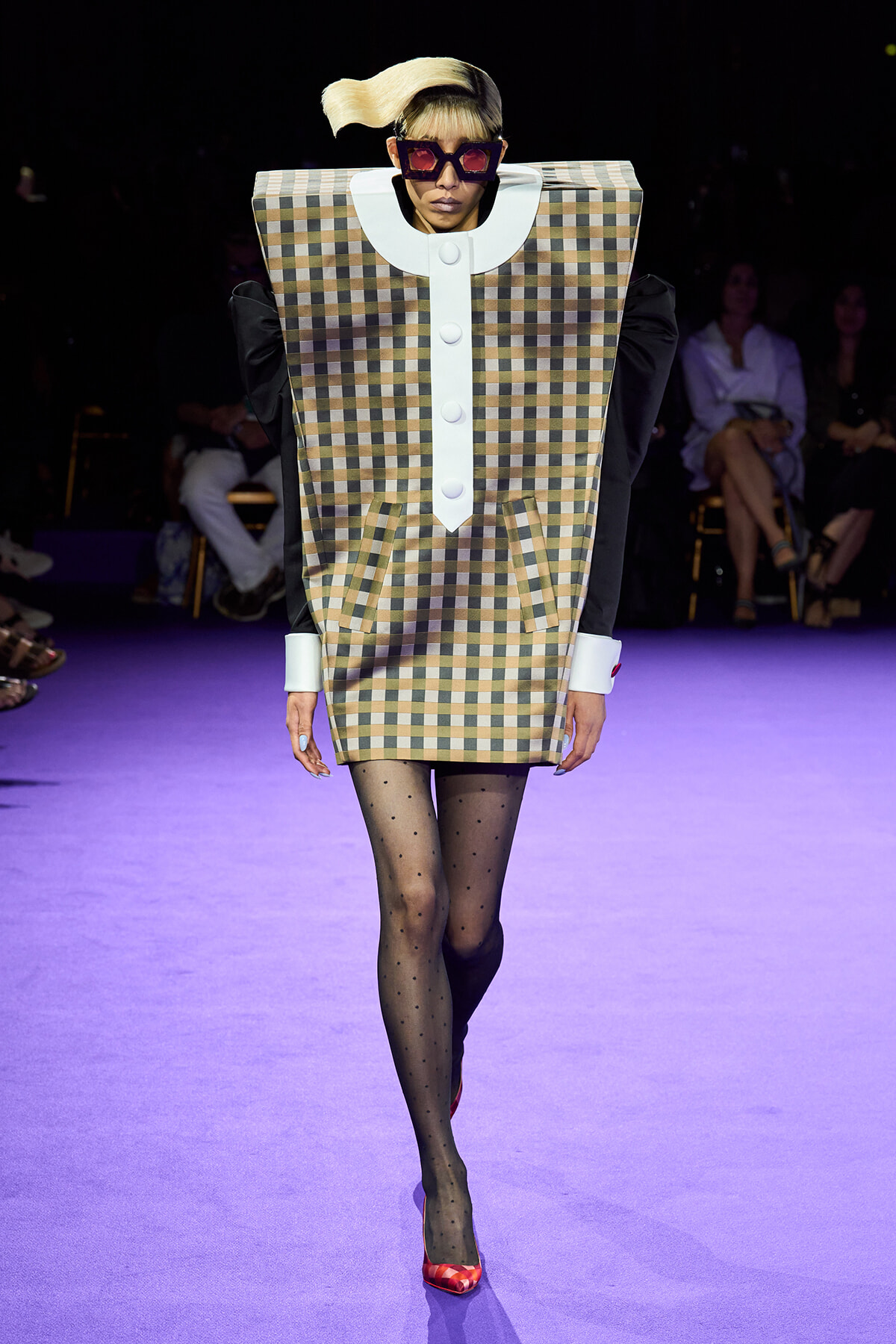 Model walks the runway in an oversized plaid tunic with white trim and buttons, black sleeves, patterned tights, and red heels under purple lighting.