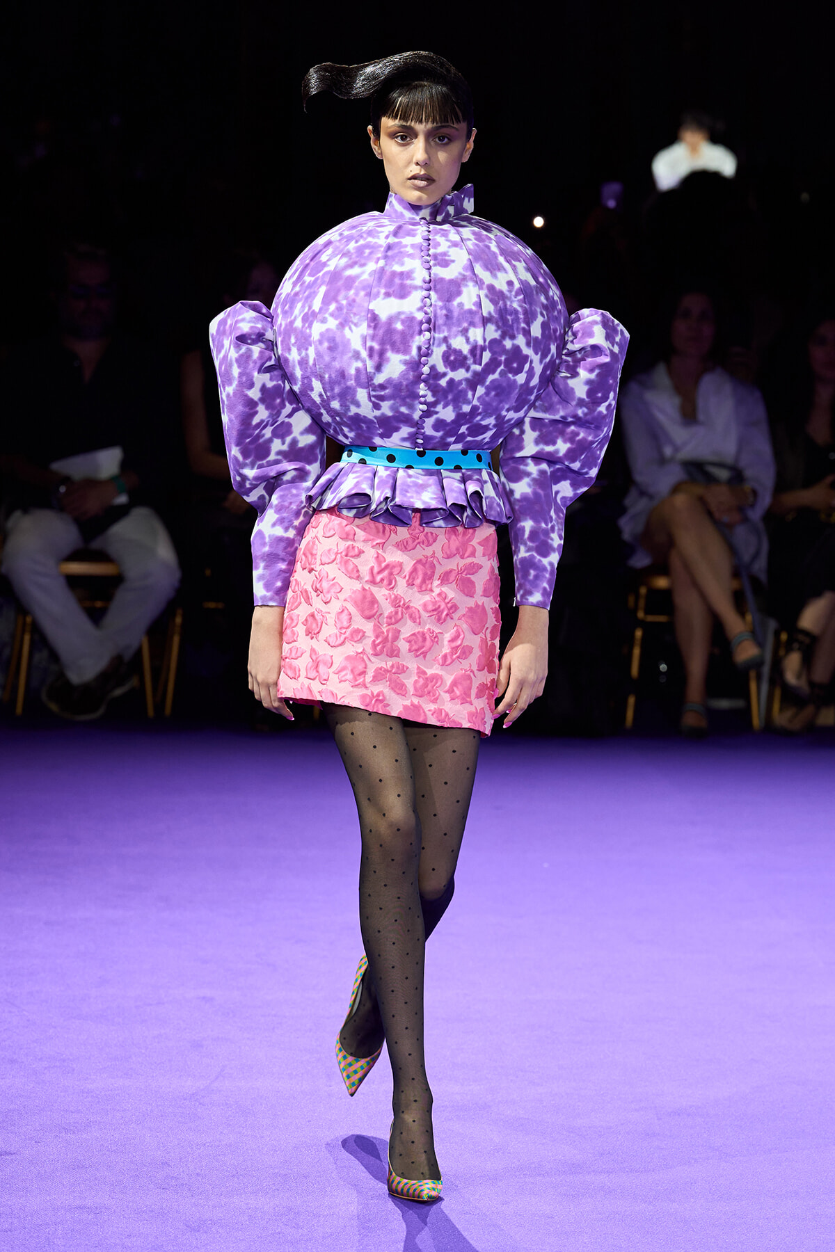 Model walking a runway in a purple tie-dye puff-sleeve top with a pink floral mini skirt and colorful checkered heels.
