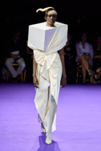 Model walks the runway in a white sculptural gown with a large cube shoulderpiece and a wrapped bow at the waist.