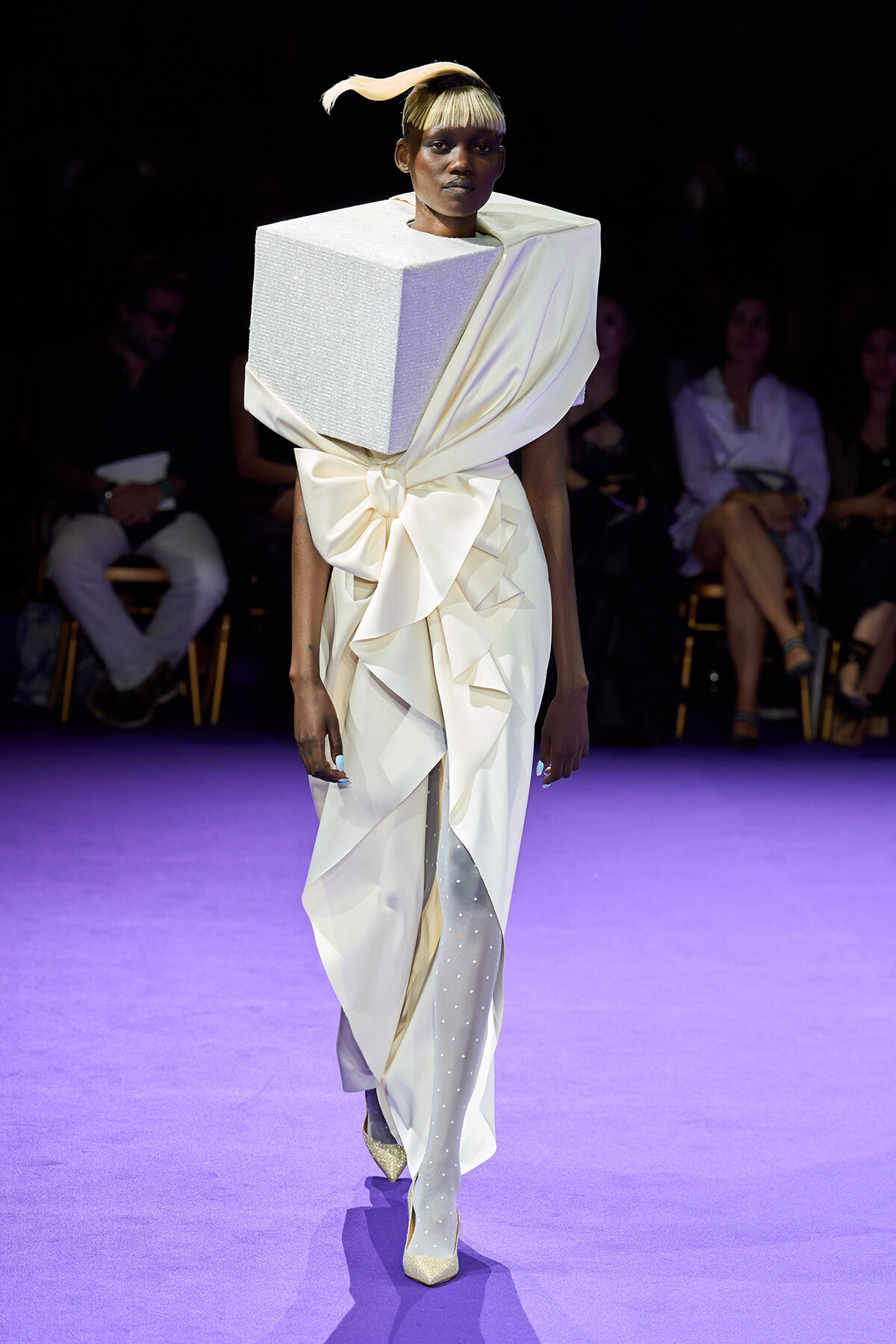 Model walks the runway in a white sculptural gown with a large cube shoulderpiece and a wrapped bow at the waist.