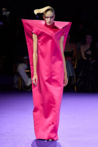 Model on a runway wearing a hot pink satin gown with angular cape-like shoulders and a high collar.
