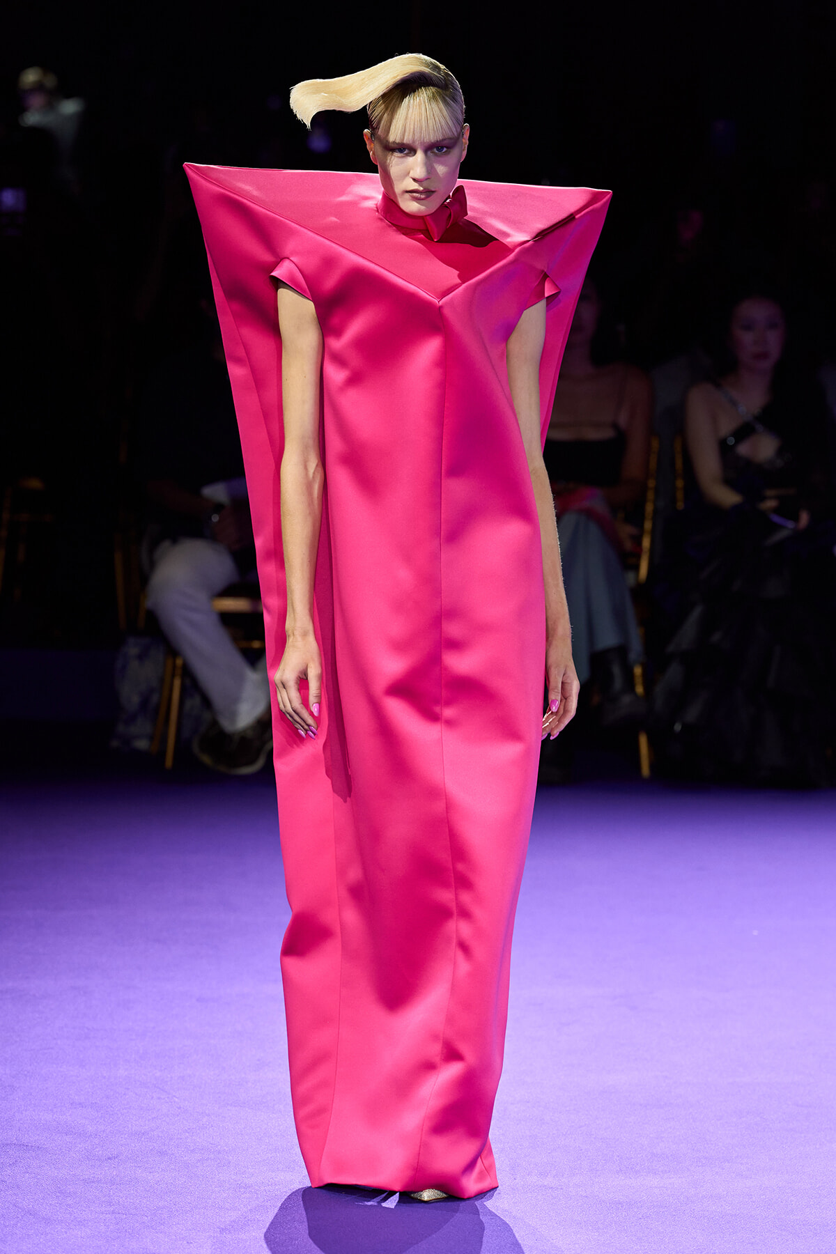 Model on a runway wearing a hot pink satin gown with angular cape-like shoulders and a high collar.