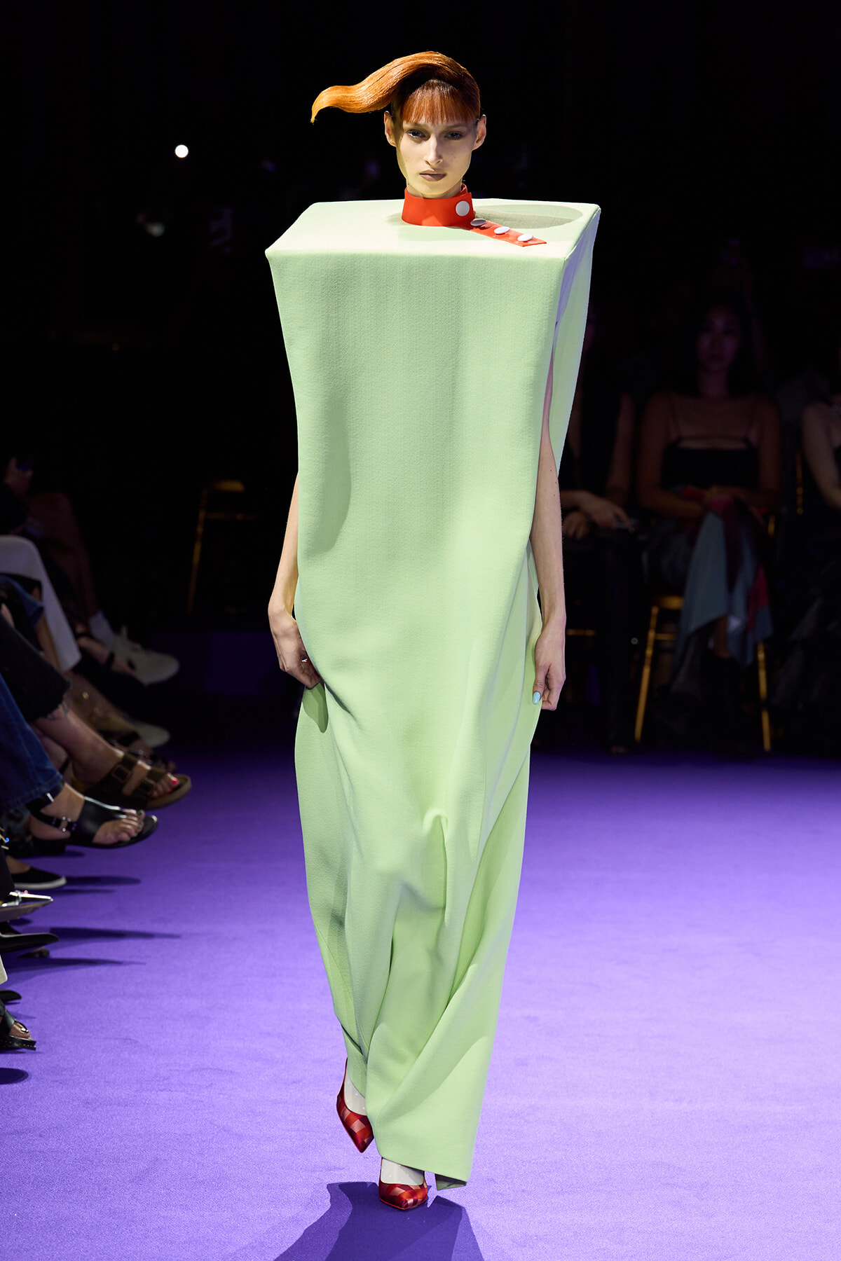 Fashion model walking a runway in a mint-green, boxy, sleeveless gown with a red collar and red-and-white accessories; bold orange hairstyle enhances the look, on a purple-lit catwalk.