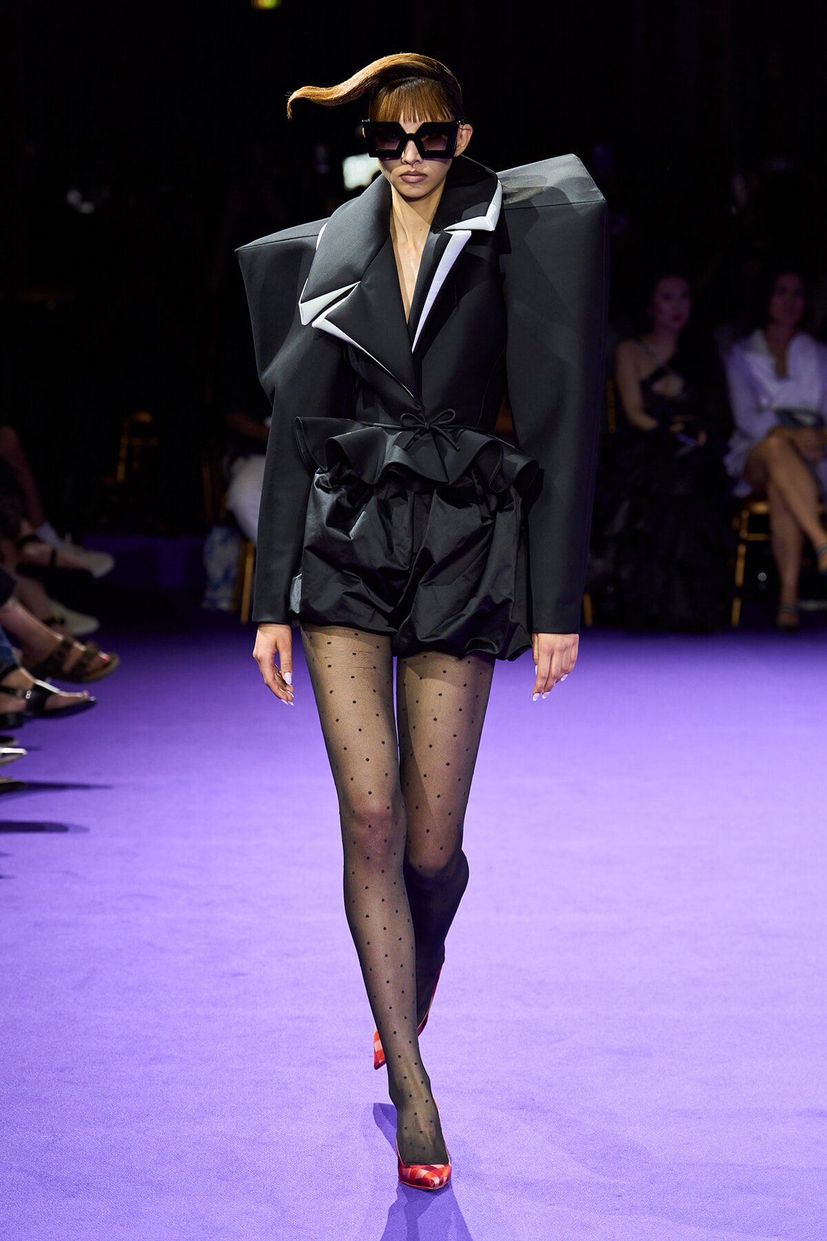 Runway model in an avant-garde black ensemble with oversized shoulders, sculpted waist, sheer polka-dot tights, and red heels.