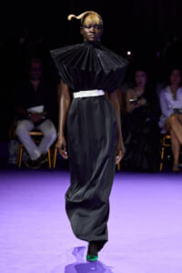 Model walks the runway in a black sculptural gown with a voluminous pleated cape collar, white waist belt, and a long satin skirt; neon green shoes add a pop of color.
