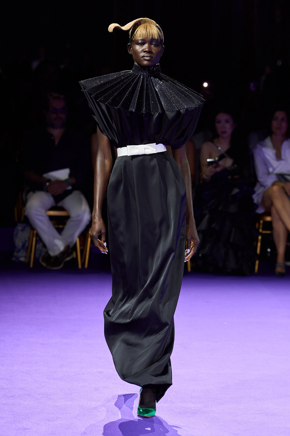 Model walks the runway in a black sculptural gown with a voluminous pleated cape collar, white waist belt, and a long satin skirt; neon green shoes add a pop of color.