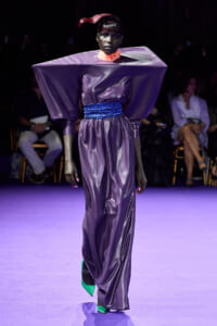 Model walking a fashion runway in a shiny purple gown with dramatic square-shoulder sleeves and a blue patterned belt. Pink neck scarf and emerald-green shoes complete the look.