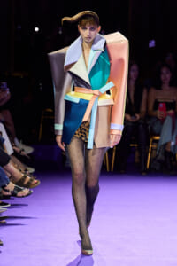 Model walks the runway in a oversized color-block trench with pastel belt, teal tights, and polka-dot stockings on a purple-lit catwalk.
