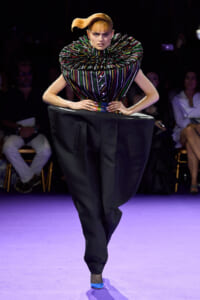 Model on a purple runway wearing a multicolored striped sculptural top with a large ruffled collar and oversized black trousers, hands on hips.