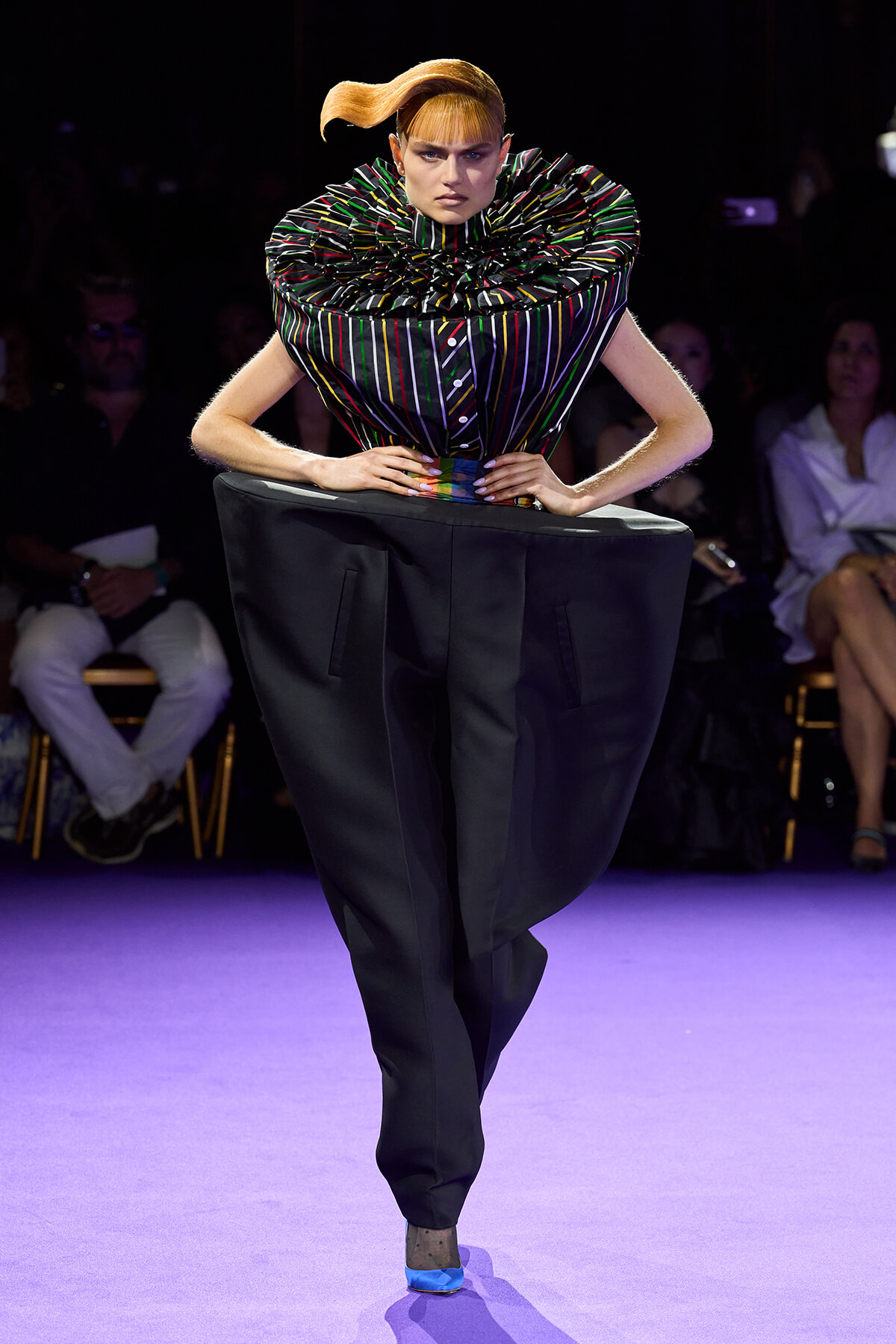 Model on a purple runway wearing a multicolored striped sculptural top with a large ruffled collar and oversized black trousers, hands on hips.