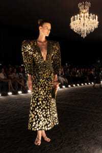 Model wearing a long black gown with gold sequin pattern walks the runway under a chandelier, audience watching in the background.