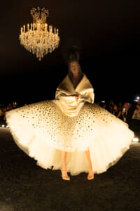 Model on a runway wearing a cream, voluminous gown with a dotted tulle skirt beneath a hanging chandelier.