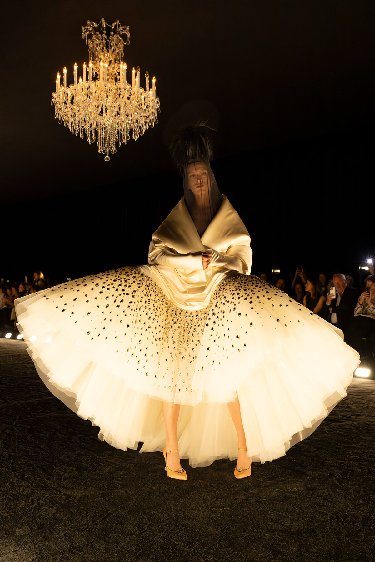 Model on a runway wearing a cream, voluminous gown with a dotted tulle skirt beneath a hanging chandelier.