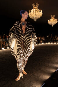 Model walks a zebra-striped cape over bare torso with blue head wrap and oversized blue pendant on a fashion runway under chandeliers.