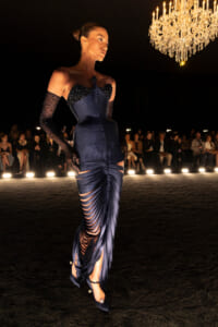 Model walks the runway in a navy, ruched gown with cutouts and beaded bodice, under a chandeliers glow.