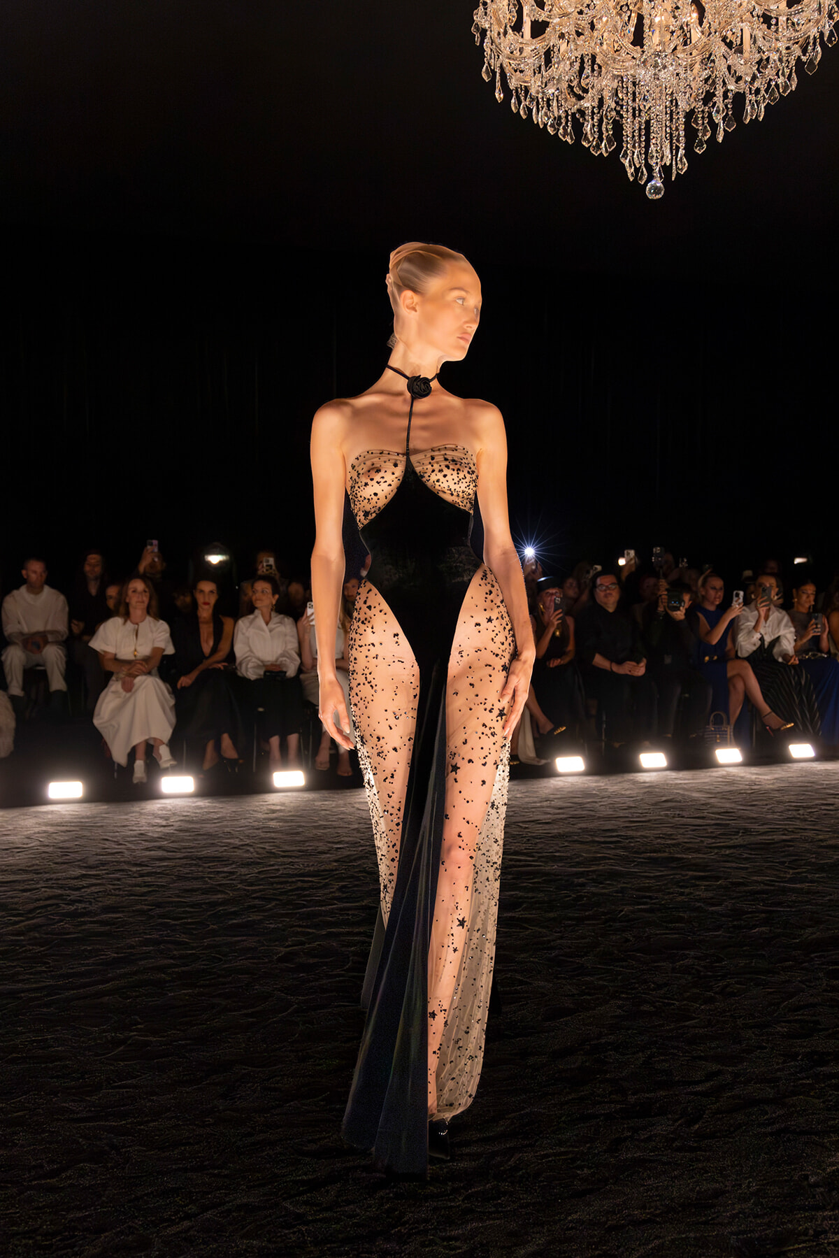 Model on a runway wearing a black sheer gown with crystal embellishments under a chandelier, audience in the background men and women watching.
