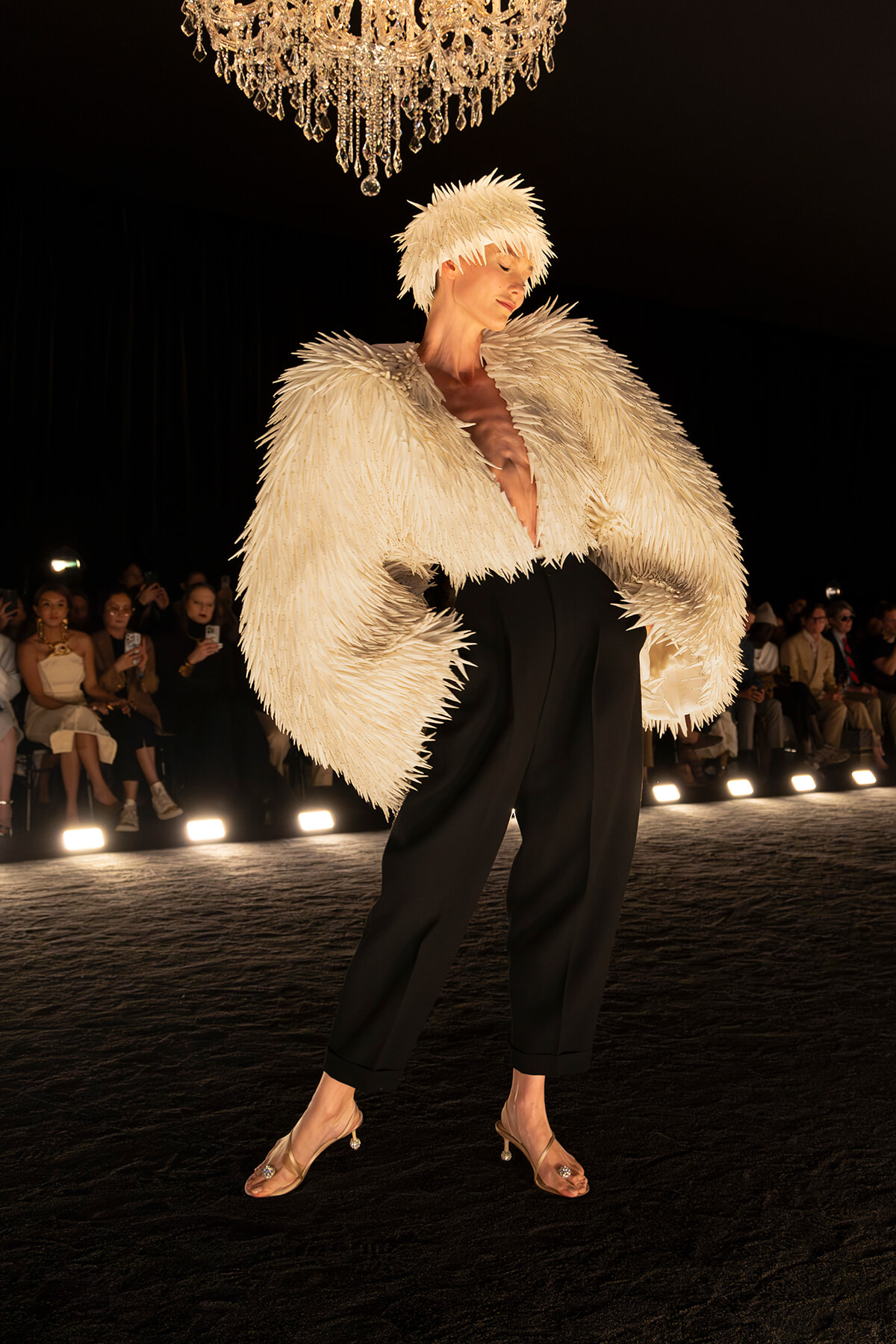 Model on a runway wearing a dramatic white feathered coat, black pants, and nude heels, with a chandelier overhead and spectators in the background.