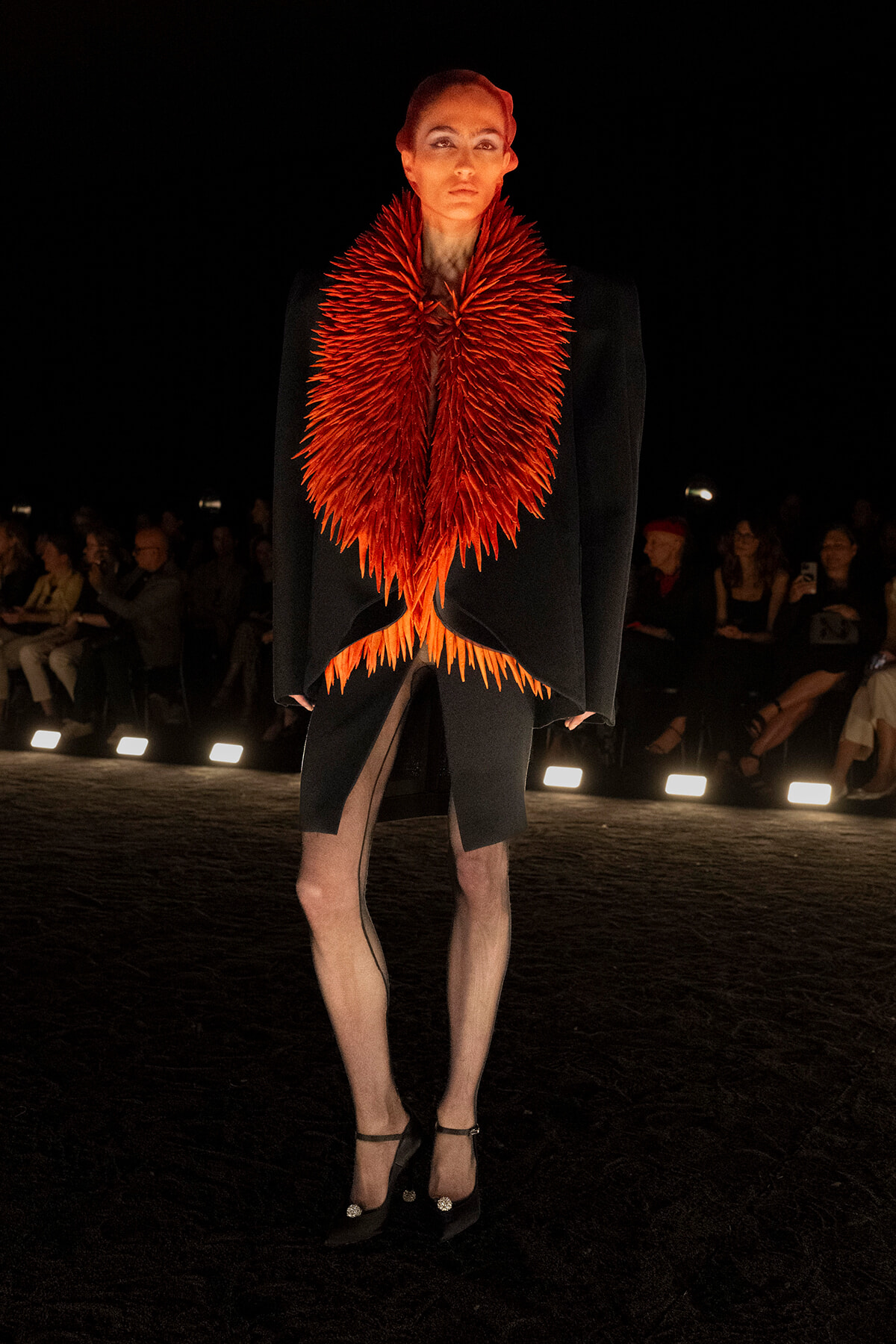 Model on a dark runway wearing a black avant-garde coat with a dramatic red feather collar, audience in the background.