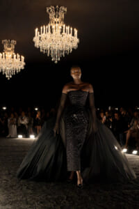 Model walks a dark runway in a strapless, sequined black gown with a dramatic cape, chandeliers overhead.