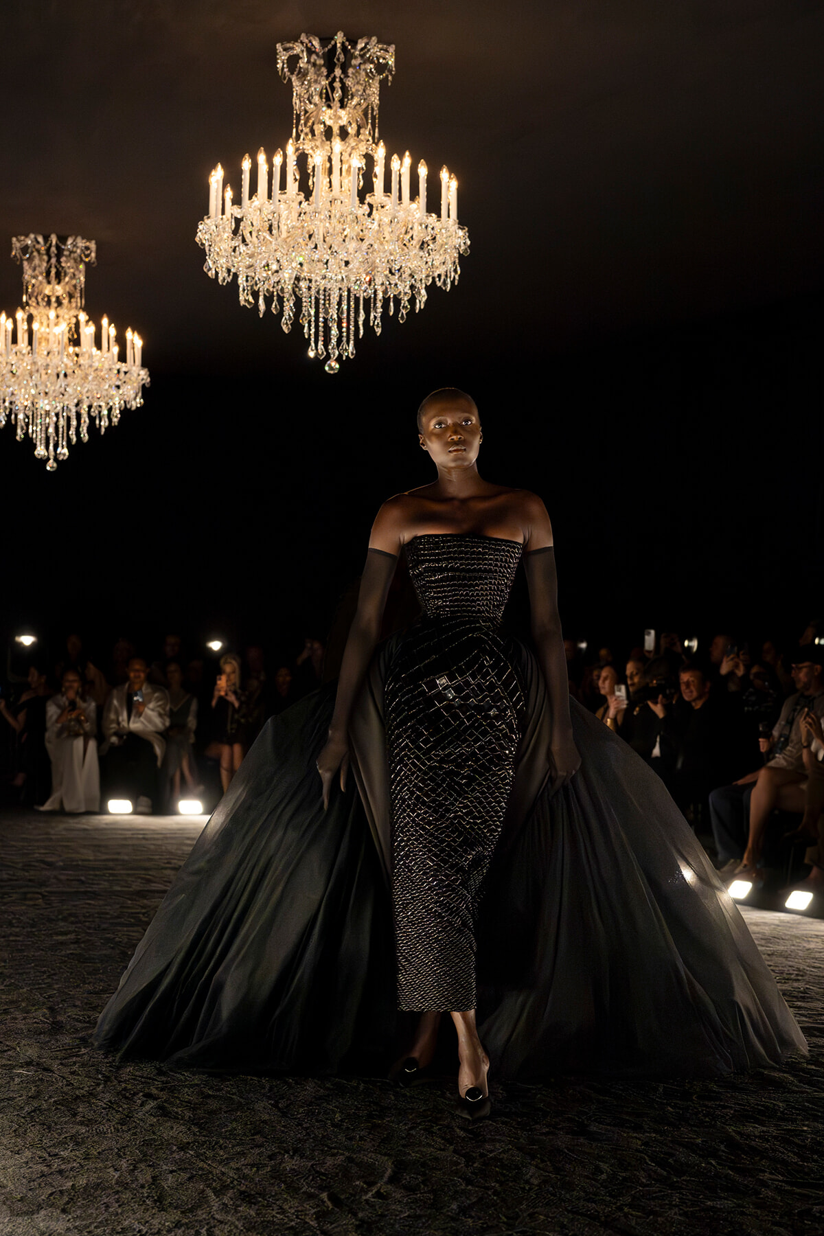 Model walks a dark runway in a strapless, sequined black gown with a dramatic cape, chandeliers overhead.