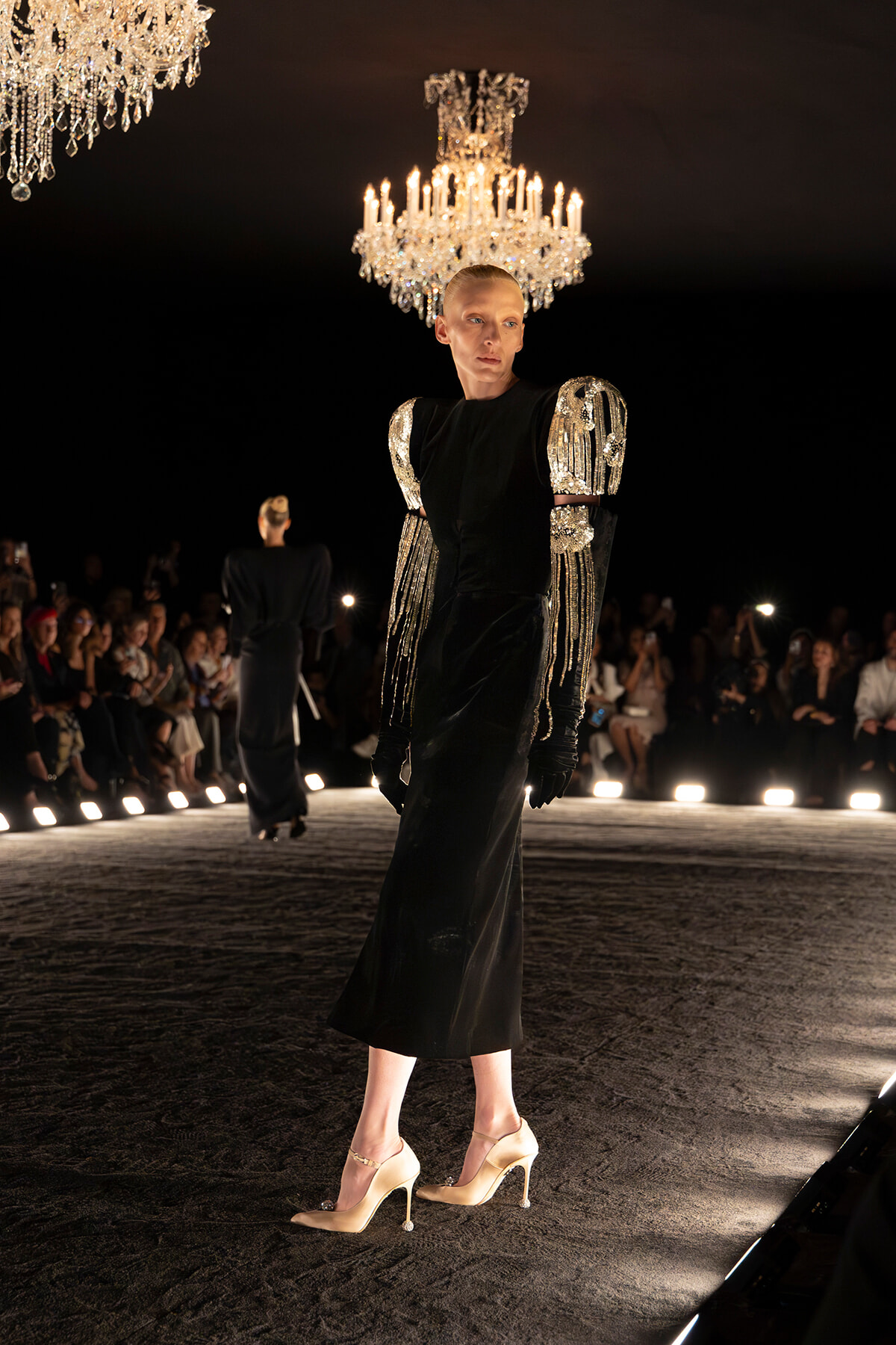 Model on a runway wearing a black gown with silver beaded shoulder embellishments and fringe, under chandeliers.