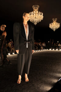 A male model on a dark runway wearing a tailored black blazer with gold trim, white inner lapels, and cropped trousers, standing under crystal chandeliers as an audience observes in the background.
