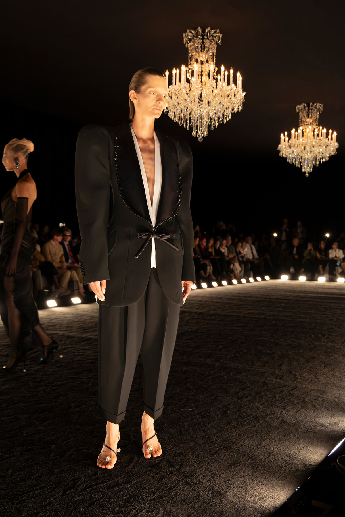 A male model on a dark runway wearing a tailored black blazer with gold trim, white inner lapels, and cropped trousers, standing under crystal chandeliers as an audience observes in the background.