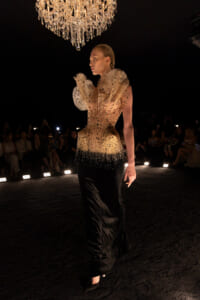 Model on a runway wearing a gradient gold-to-black gown, dramatic ruffled shoulders, under a crystal chandelier.