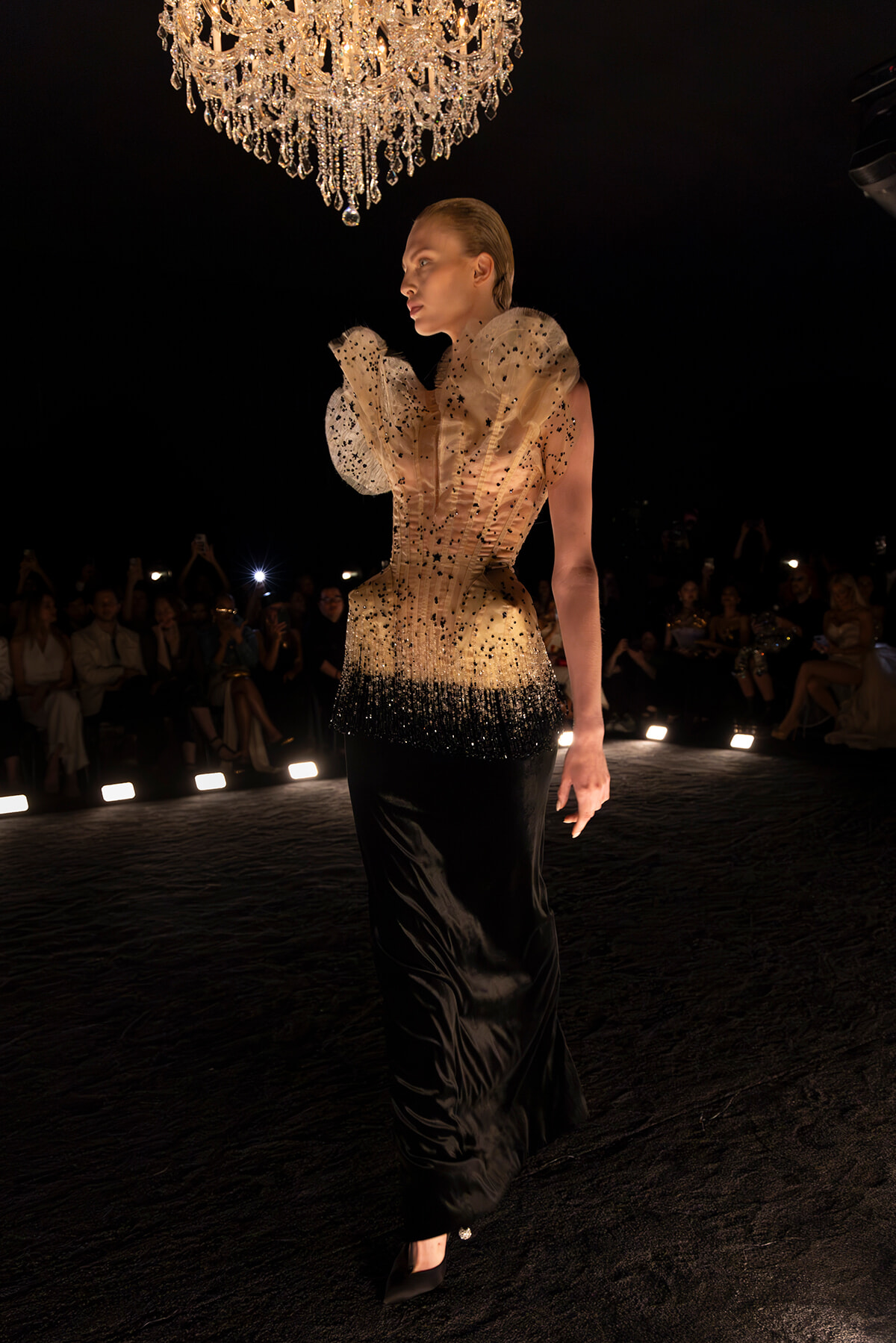 Model on a runway wearing a gradient gold-to-black gown, dramatic ruffled shoulders, under a crystal chandelier.
