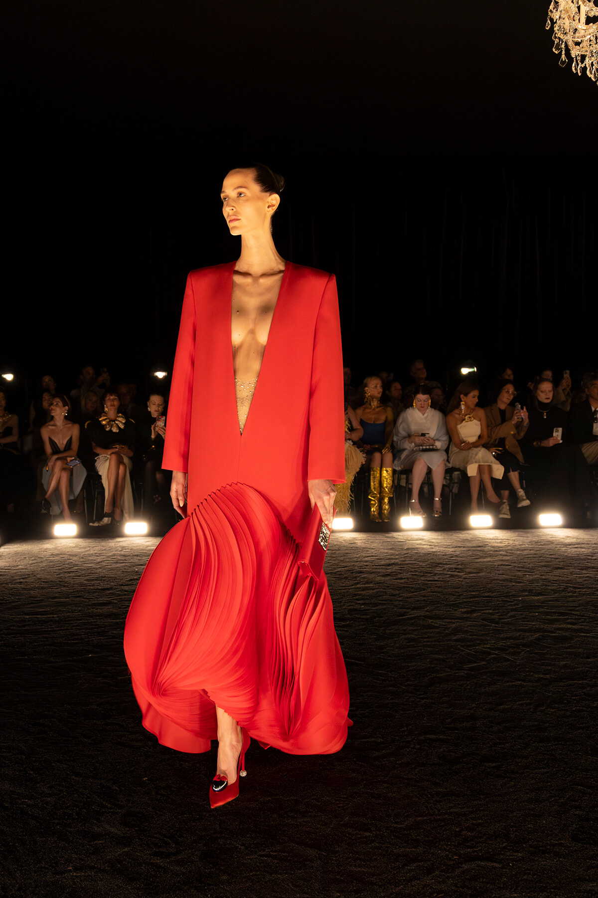 Model walks a runway in a bold red outfit: a structured blazer with a deep V and a flowing pleated red skirt; audience sits in the background under dim lights.