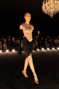 Model walks a runway in a nude spiked bodysuit with a black ruched skirt and long black gloves, chandelier overhead.