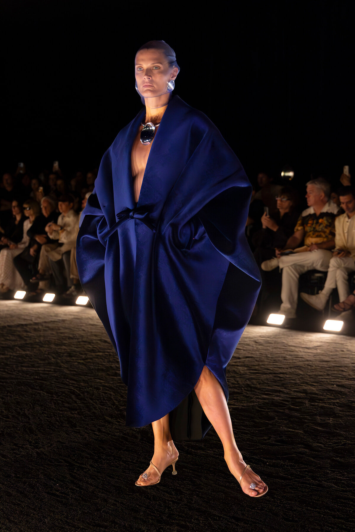 Model walking a runway in a deep blue oversized satin robe with a waist belt, bold necklace, and nude heeled sandals; dark audience backdrop.