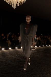 Model walks a runway in a dark, textured gown with oversized shoulders and a belt, holding a round metallic clutch.