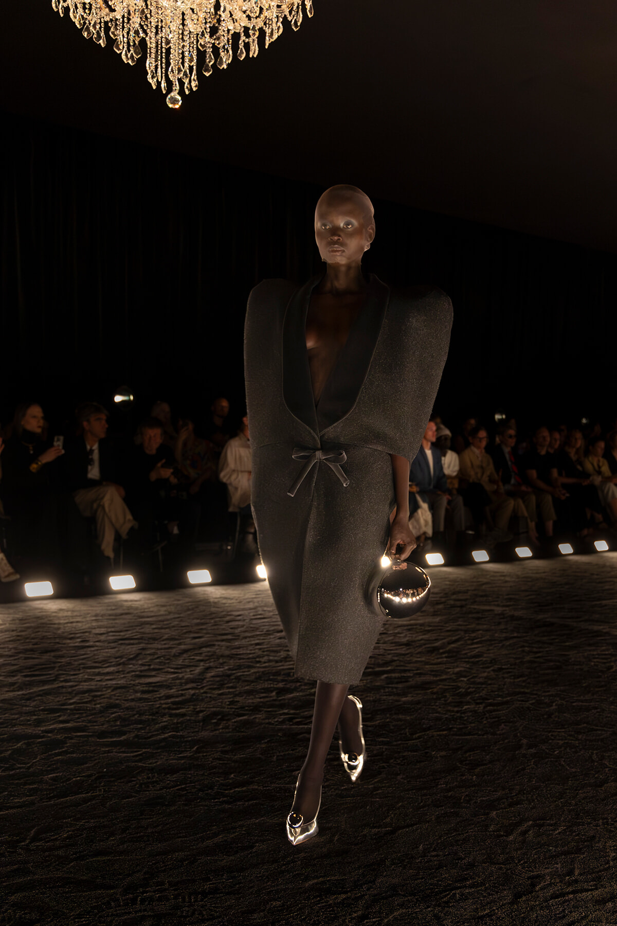 Model walks a runway in a dark, textured gown with oversized shoulders and a belt, holding a round metallic clutch.